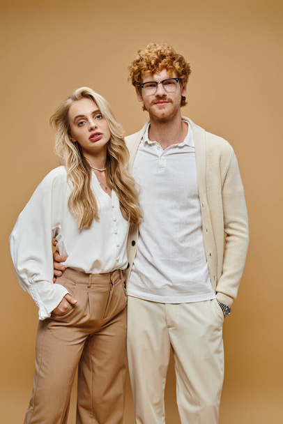 trendy young couple in light-colored clothes with hands in pockets looking at camera on beige - Photo, Image