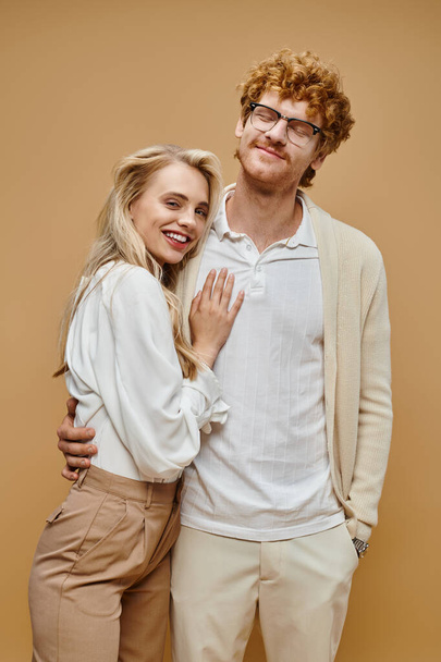 cheerful blonde woman embracing trendy redhead man and looking at camera on beige, classic style - Photo, Image