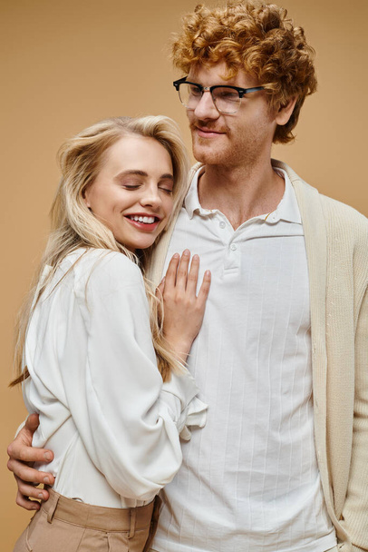 gelukkig roodharige man in bril omarmen stijlvolle blonde vrouw glimlachen met gesloten ogen op beige - Foto, afbeelding