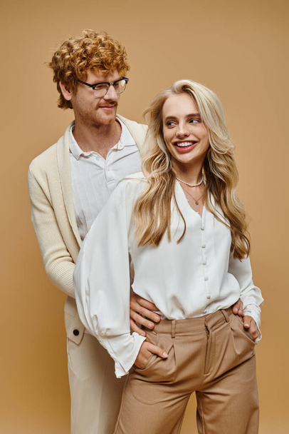 rothaariger Mann mit Brille umarmt blonde Frau, die mit Händen in Taschen auf beige steht, Mode - Foto, Bild
