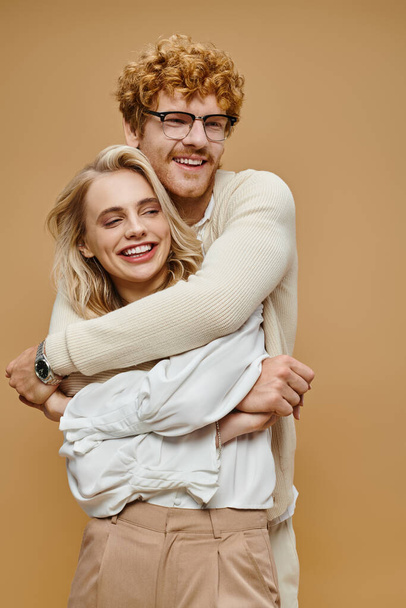 šťastný ryšavý muž v brýlích objímající trendy blondýny žena na béžové, staré peníze styl pár - Fotografie, Obrázek