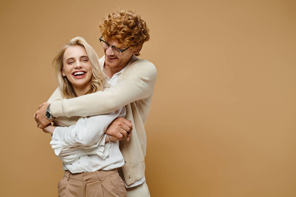glimlachende roodharige man in bril knuffelen blonde stijlvolle vrouw lachen op beige, oud geld stijl - Foto, afbeelding