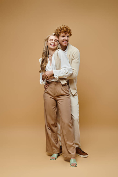 повна довжина радісної молодої пари в старому грошовому стилі повсякденний одяг, що обіймається на бежевому тлі - Фото, зображення