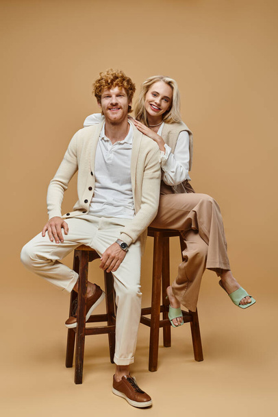 alegre jovem casal em roupas elegantes sentado em cadeiras e olhando para a câmera no fundo bege - Foto, Imagem
