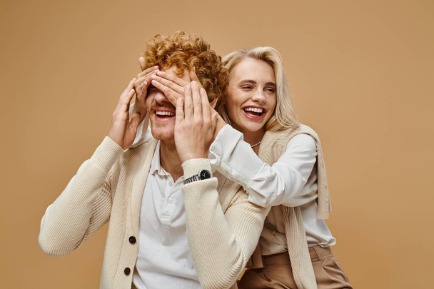 playful blonde woman covering eyes of stylish redhead man on beige, old money style fashion - Photo, Image