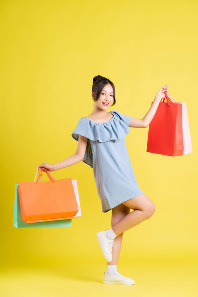 wizerunek pięknej azjatyckiej dziewczyny w sukience trzymającej torbę na zakupy w dłoni pozującej na żółtym tle - Zdjęcie, obraz