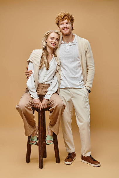 full length of redhead man embracing blonde woman sitting on chair and looking at camera on beige - Photo, Image