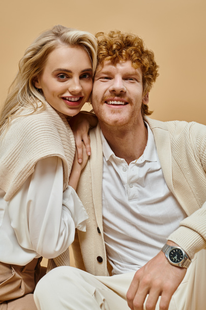 pleased blonde woman and redhead man in light-colored outfit looking at camera on beige backdrop - Photo, Image