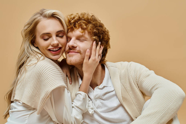 delighted blonde woman with closed eyes embracing head of stylish redhead man on beige backdrop - Photo, Image