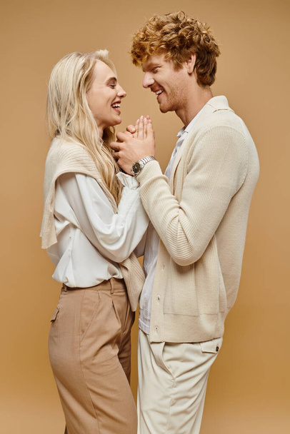 vista lateral do casal na moda roupas de cor clara de mãos dadas e rindo em pano de fundo bege - Foto, Imagem