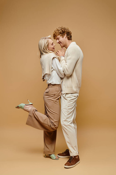 full length of couple in stylish light-colored clothes holding hands and laughing on beige backdrop - Photo, Image