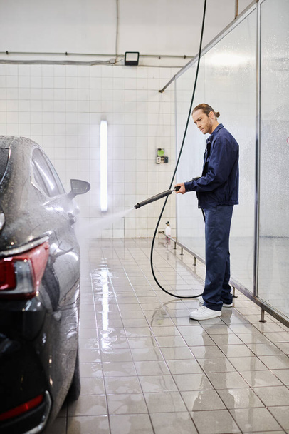 atraente trabalhador masculino profissional em azul uniforme lavagem carro preto com mangueira enquanto na garagem - Foto, Imagem