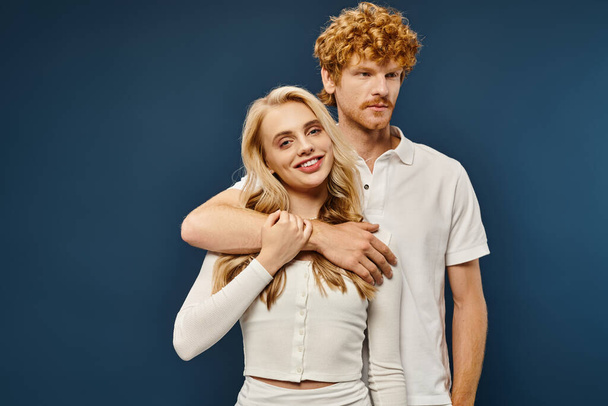 handsome redhead man embracing cheerful blonde woman in white stylish clothes on blue backdrop - Photo, Image