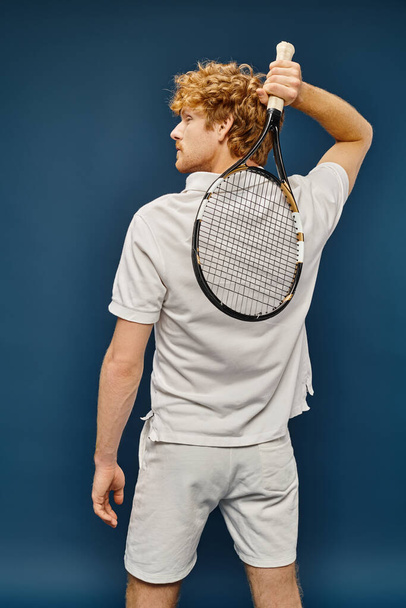 terug uitzicht op jonge roodharige man in wit tennis outfit poseren met racket op blauwe, tijdloze mode - Foto, afbeelding