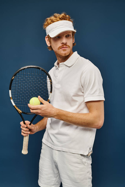 sportieve man in wit tennis outfit en zonneklep met racket en bal op blauwe, rijke sport - Foto, afbeelding