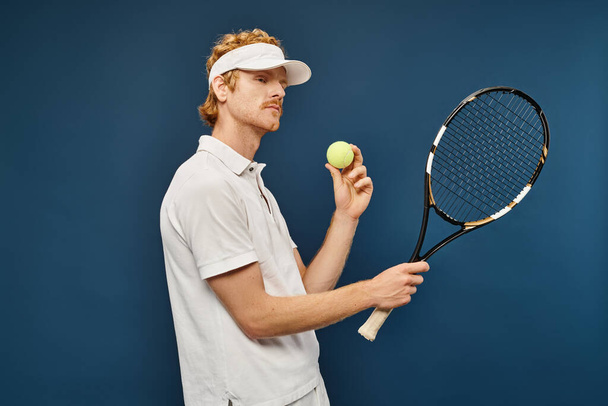 Молодой красивый рыжий мужчина в белой одежде и солнцезащитном козырьке держит ракетку и теннисный мяч на голубом - Фото, изображение