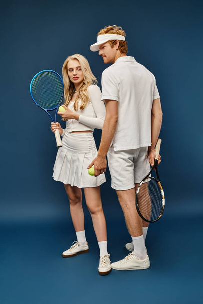 longitud completa de la pareja joven en traje de tenis blanco con raquetas y bolas en azul, estilo de dinero viejo - Foto, imagen