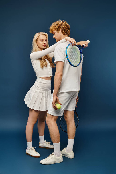 stylish blonde woman with tennis racquet embracing redhead man in white attire on blue, full length - Photo, Image