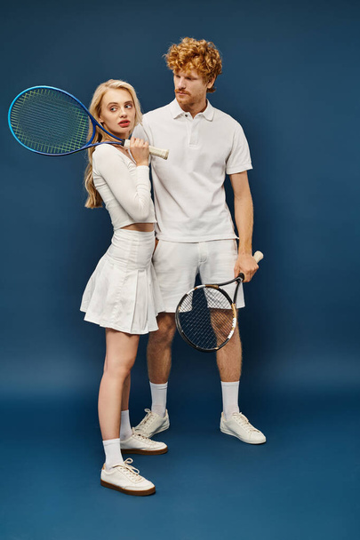 longitud completa de pelirrojo hombre mirando a la mujer rubia con raqueta de tenis mirando hacia otro lado en azul - Foto, imagen