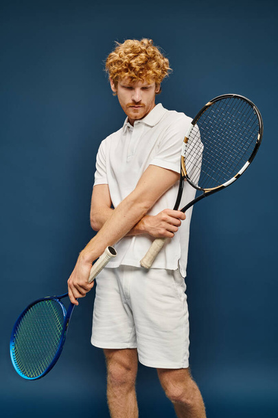 arystokratyczna ruda mężczyzna w białym stroju pozowanie z rakiety tenisowe pozowanie na niebieskim tle - Zdjęcie, obraz