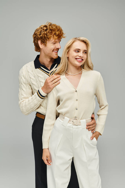 happy redhead man in casual attire embracing young blonde woman on grey backdrop, fashionable couple - Photo, Image
