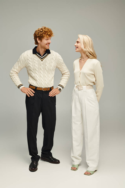 аристократичный рыжий мужчина с руками на бедрах смотрит на блондинку в модной одежде на сером - Фото, изображение