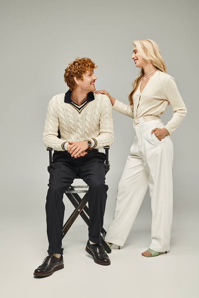 модная блондинка улыбается рядом с молодым рыжим мужчиной, сидящим на стуле на серой, классической моде - Фото, изображение