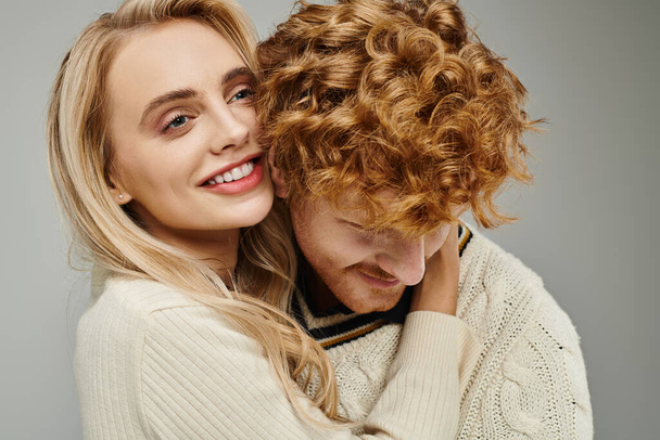 happy and fashionable blonde woman embracing young redhead man on grey, trendy couple relationship - Photo, Image