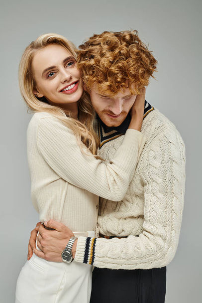 uśmiechnięta i stylowa blondynka przytulająca młodego rudego mężczyznę na szarym, modnym związku para - Zdjęcie, obraz