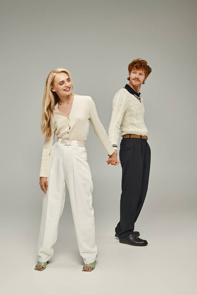 volledige lengte van gelukkig jong roodharige man en blonde vrouw houden handen op grijze achtergrond in studio - Foto, afbeelding