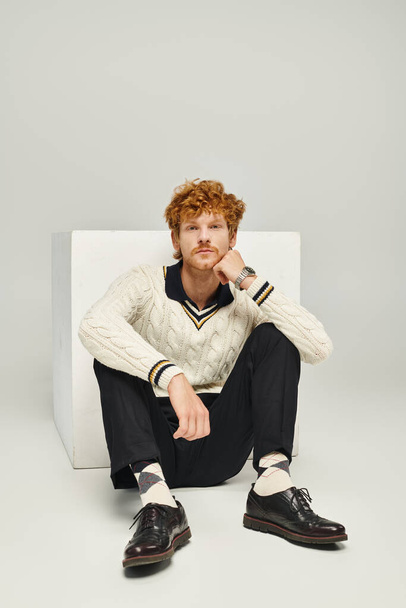 задумчивый мечтательный мужчина в повседневной одежде, смотрящий в камеру, сидящую рядом с белым кубом на сером фоне - Фото, изображение