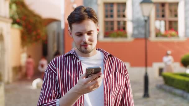 ハッピー笑顔のブルネットひげの男は,インターネットソーシャルメディアWebアプリを閲覧するテキストメッセージを入力するスマートフォンを使用して,オンラインチャット. 市街地のカメラを眺めている男. ライフスタイル - 映像、動画