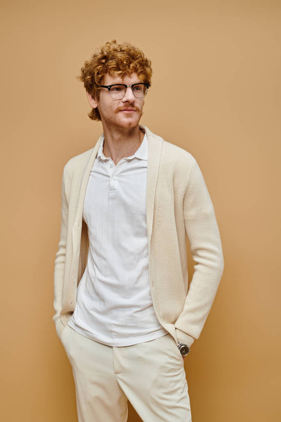 glimlachende elegante roodharige man in bril die wegkijkt terwijl hij met handen in zakken op beige staat - Foto, afbeelding