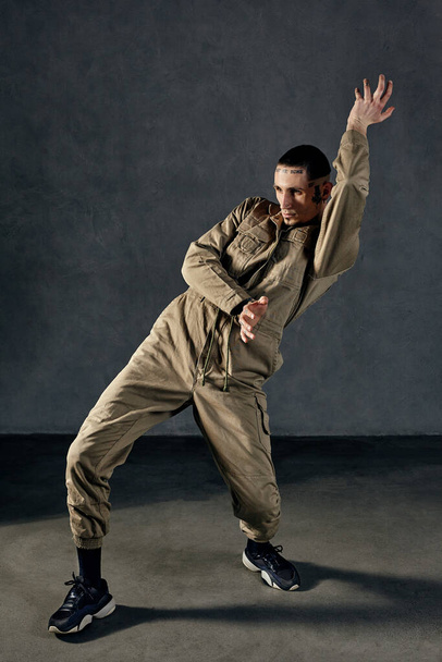 Young stately guy with tattooed body and face, earrings, beard. Dressed in khaki overalls and black sneakers. He is dancing against gray studio background. Dancehall, hip-hop. Full length, copy space - Photo, Image