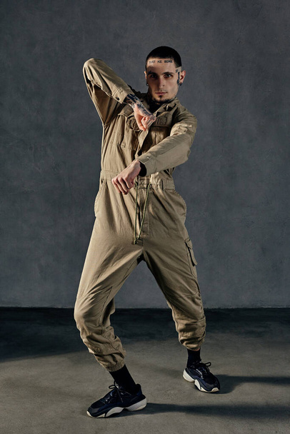 Young active man with tattooed body and face, earrings, beard. Dressed in khaki overalls and black sneakers. He is dancing against gray studio background. Dancehall, hip-hop. Full length, copy space - Photo, Image