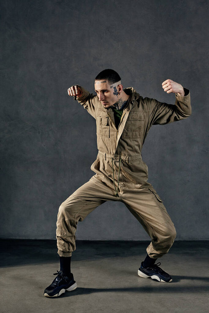 Young active male with tattooed body and face, earrings, beard. Dressed in khaki jumpsuit and black sneakers. He is dancing against gray studio background. Dancehall, hip-hop. Full length, copy space - Foto, immagini