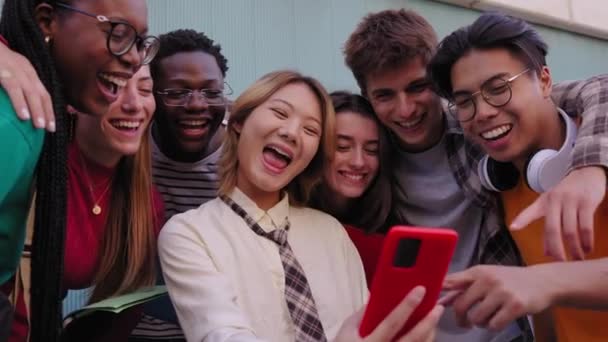 Grupa młodych międzynarodowych studentów uśmiecha się radośnie oglądając śmieszne filmy na smartfonie trzymającym azjatycką dziewczynę. Pokolenie z przyjaciółmi radosne patrząc na telefon poza kampusem wydziału - Materiał filmowy, wideo