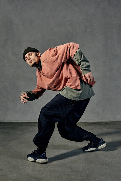 Handsome fellow with tattooed body and face, earrings, beard. Dressed in hat, colorful jumper, black pants and sneakers. Dancing on gray background. Dancehall, hip-hop. Full length, copy space - Foto, Imagen