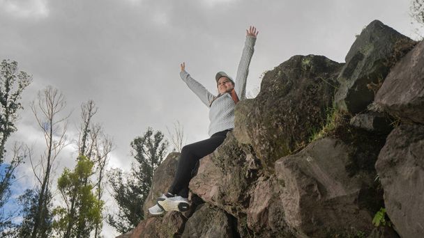 Hermosa joven latinoamericana sentada sobre piedras en la cima de una montaña con los brazos levantados victoriosamente en un día nublado - Foto, Imagen