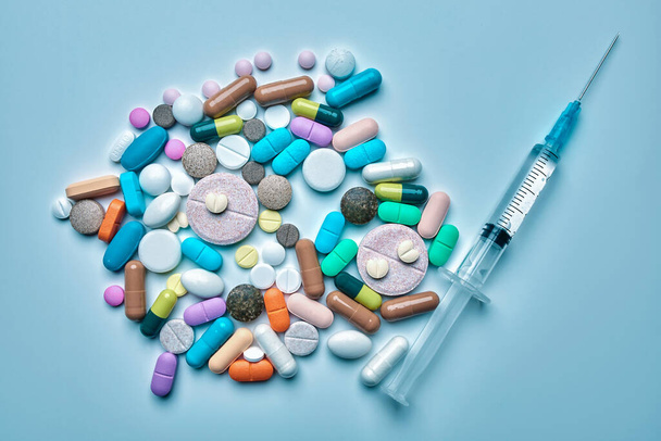 Een medische spuit met een naald en veel medicinale pillen gefotografeerd in het bovenste zicht op een lichtblauwe achtergrond, het concept van de geneeskunde en de gezondheidszorg - Foto, afbeelding