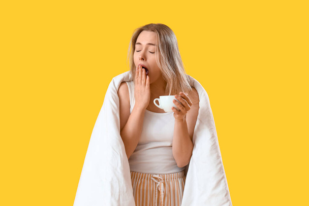 Ospalá mladá žena s dekou a šálek kávy na žlutém pozadí - Fotografie, Obrázek