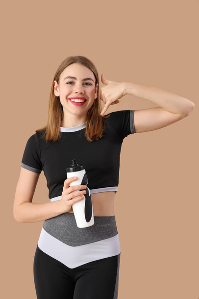 Schöne Fitnesstrainerin mit Wasserflasche zeigt "Call me" -Geste auf braunem Hintergrund - Foto, Bild