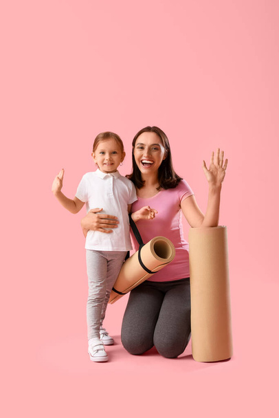 Όμορφη μητέρα και χαριτωμένη μικρή κόρη της με στρώματα γιόγκα κουνώντας τα χέρια σε ροζ φόντο - Φωτογραφία, εικόνα