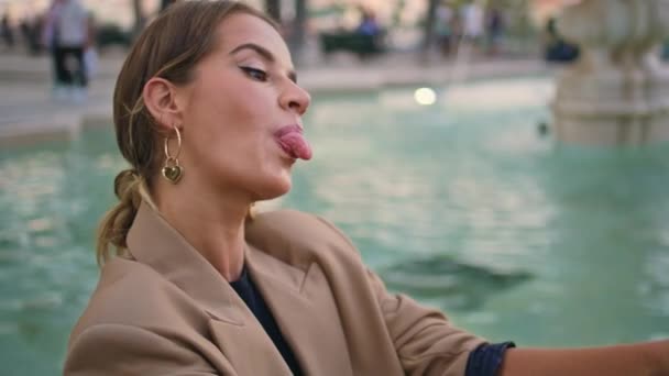 Girl taking funny selfie picture with tongue out closeup. Carefree lady texting sending social media message online at water urban fountain. Gorgeous woman making photo using cellphone in evening. - Footage, Video