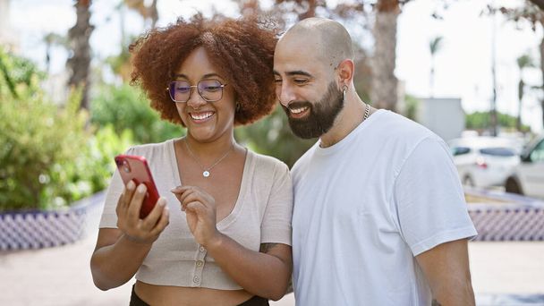 Pewna siebie, szczęśliwa międzyrasowa para uśmiechnięta razem, stojąca w słonecznym parku ze swoim smartfonem, bez wysiłku dzieląca się miłością i śmiechem na świeżym powietrzu - Zdjęcie, obraz