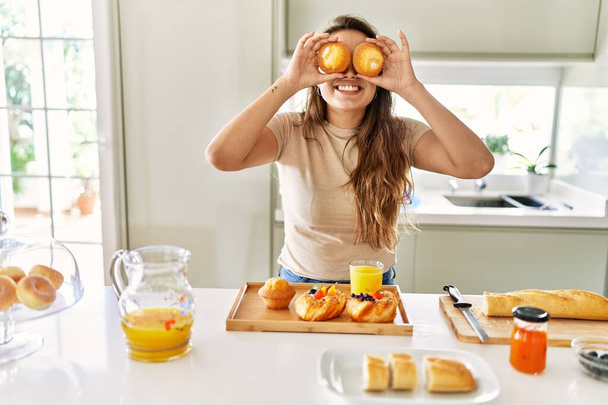 Young beautiful hispanic woman preparing breakfast holding cupcakes over eyes at the kitchen - Photo, Image
