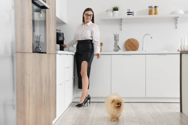 Young businesswoman with cute Pomeranian dog making coffee in kitchen - Photo, Image