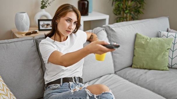 Νεαρή γυναίκα που βλέπει τηλεόραση πίνοντας καφέ στο σπίτι - Φωτογραφία, εικόνα