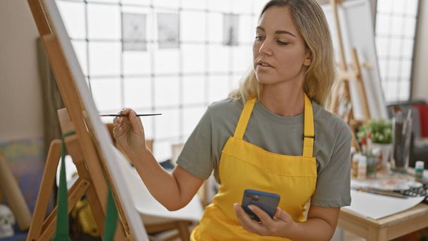 Zaostřená mladá žena umělkyně ve žluté zástěře maluje na plátno v jasném studiu, zatímco drží smartphone. - Fotografie, Obrázek
