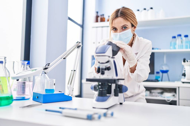 Mujer joven rubia científica usando máscara médica usando microscopio en el laboratorio - Foto, imagen